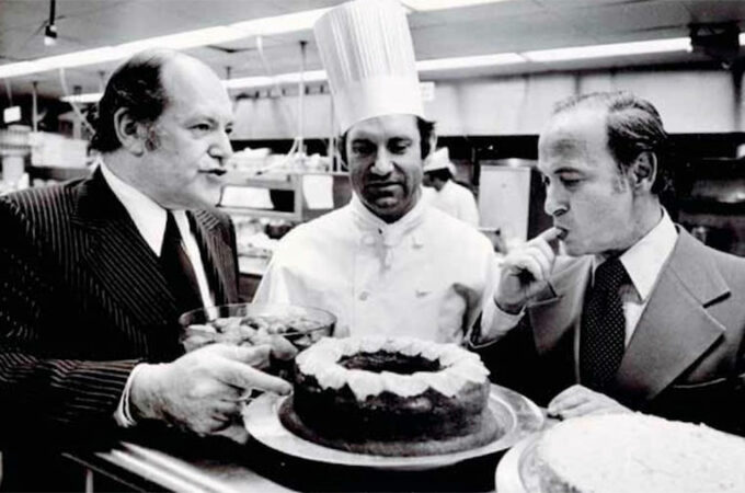 black and white photo of jow baum pointing at a freshly baked pie with chef looking on