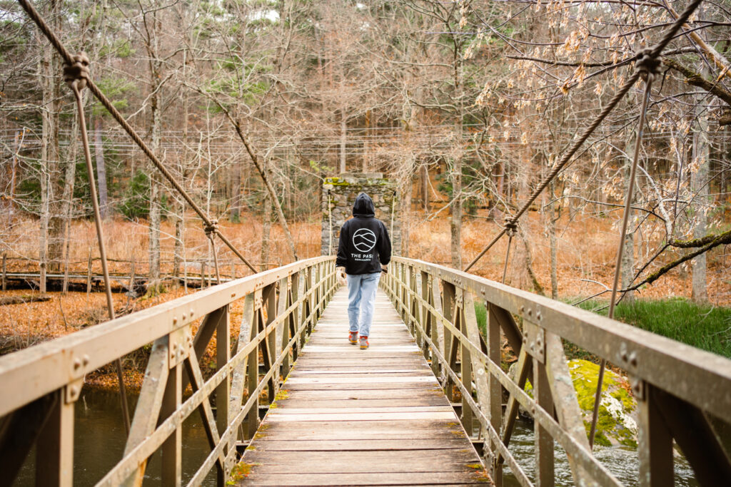 person with a hoodie walking on boardwalk over river in the woods